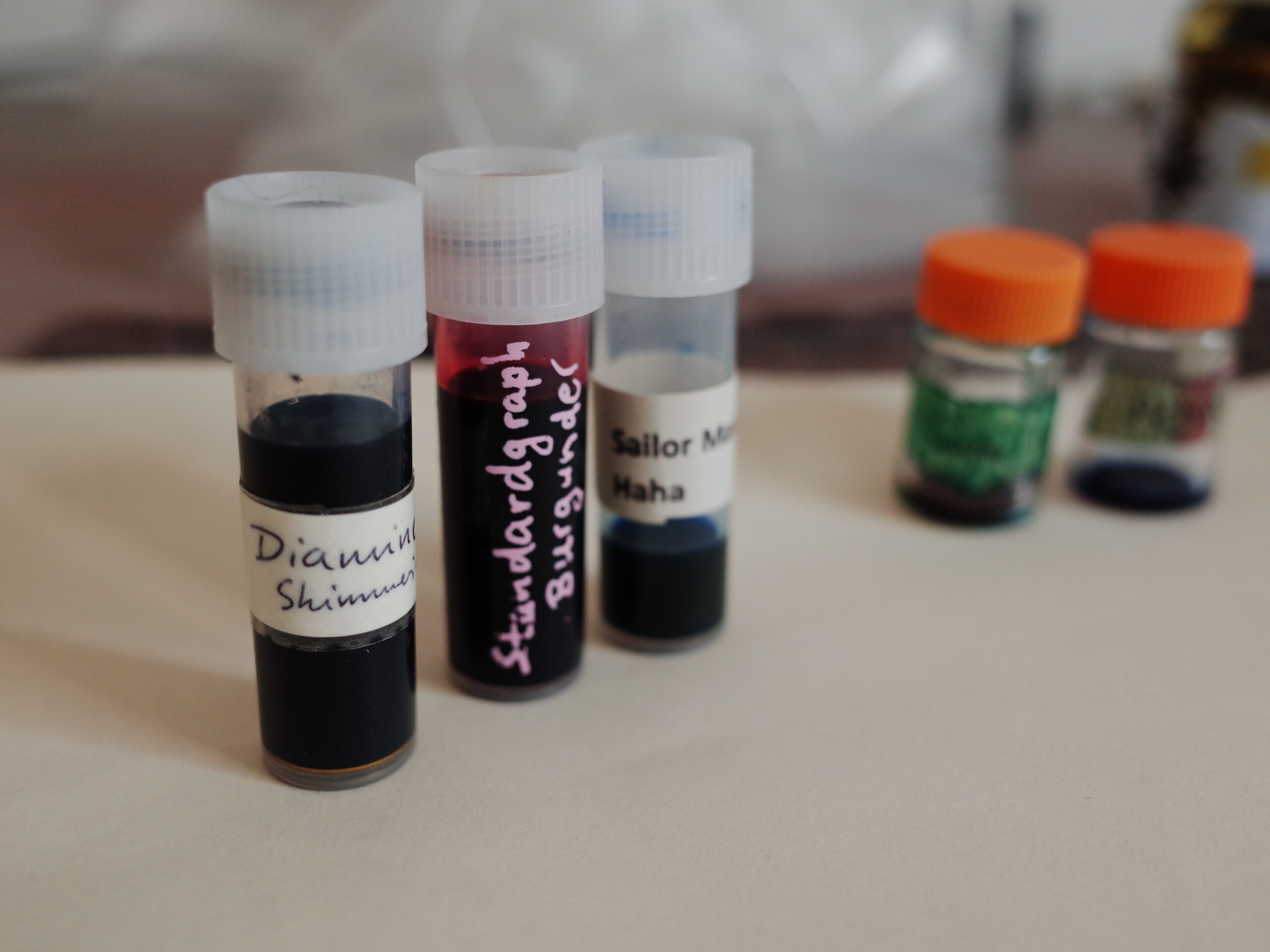 3 plastic ink sample vials with ink in it and different labels, two blurry empty sample vials in the background